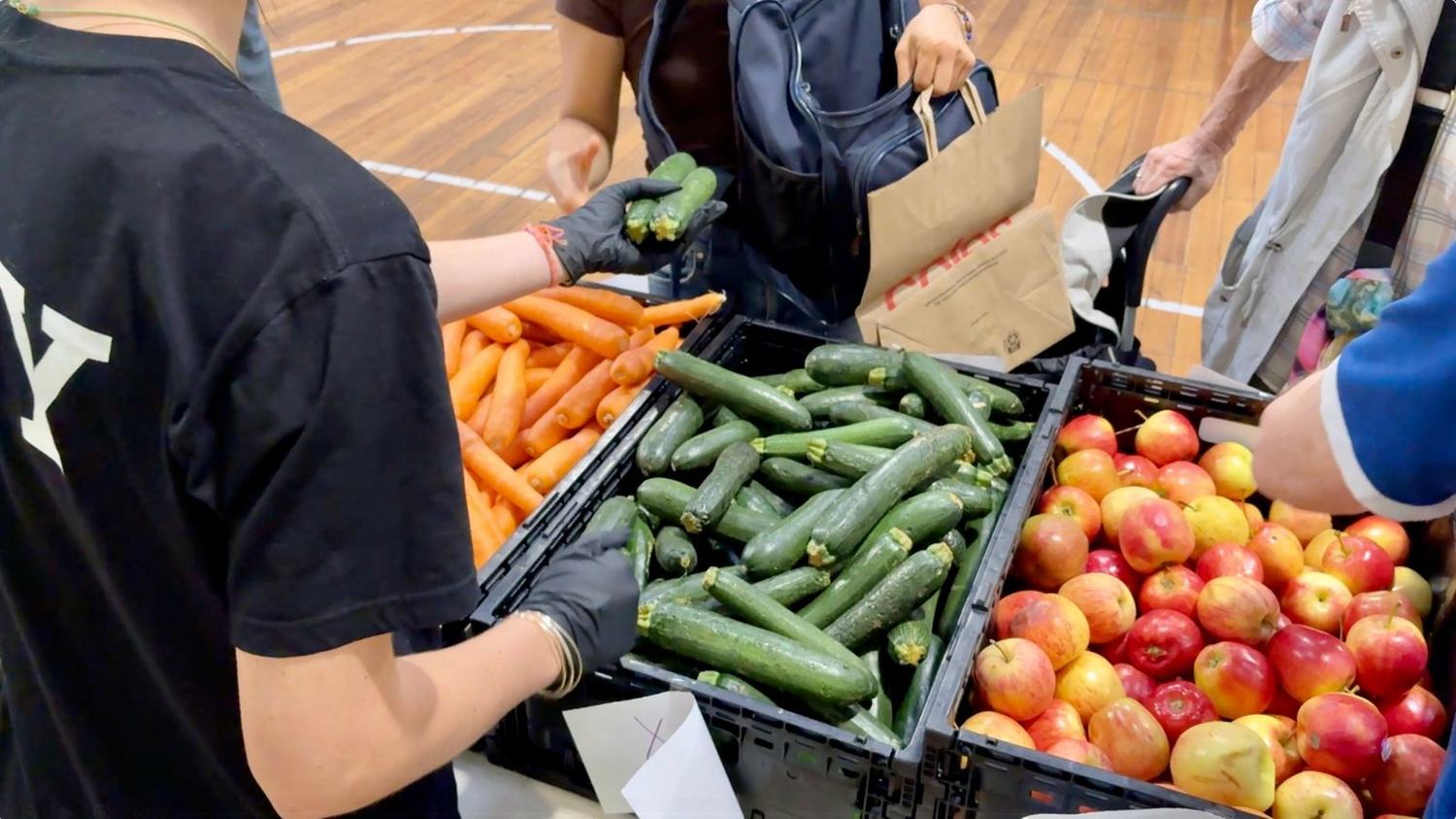 A volunteer in black gloves handing out zucchini from crates of fresh produce