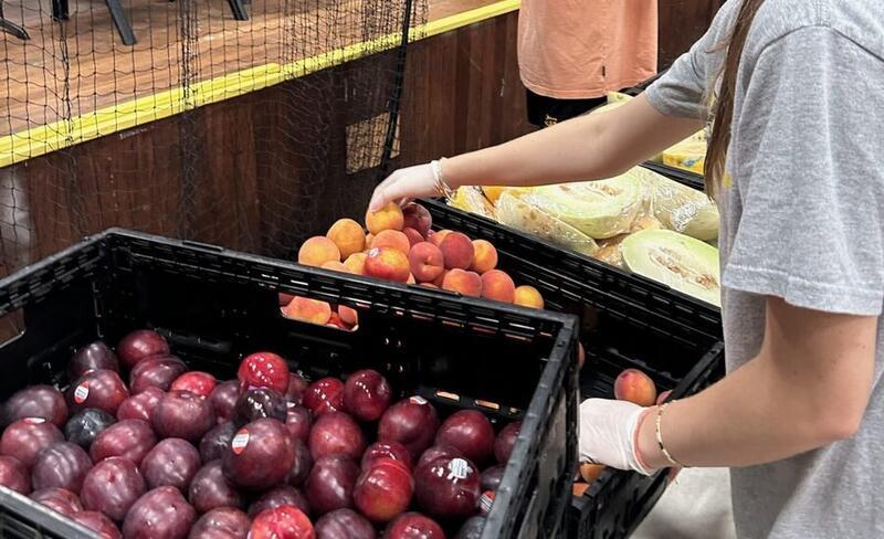 A volunteer's gloved hand picking fruit from a crate of peaches and plums