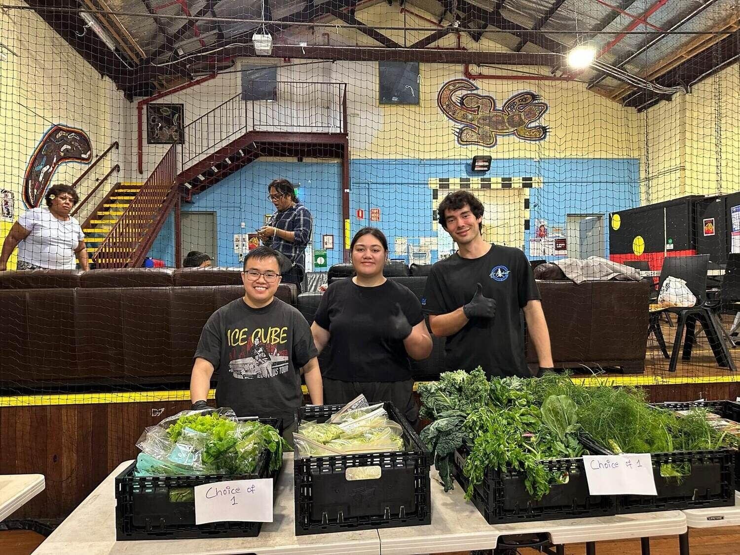 Three Food Circle volunteers giving a thumbs up in front of crates of fresh vegetables at Glebe Youth Service